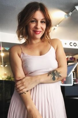 Jenn in a pale pink dress, her left arm is across her body holding her right arm. her left forearm has a tattoo.